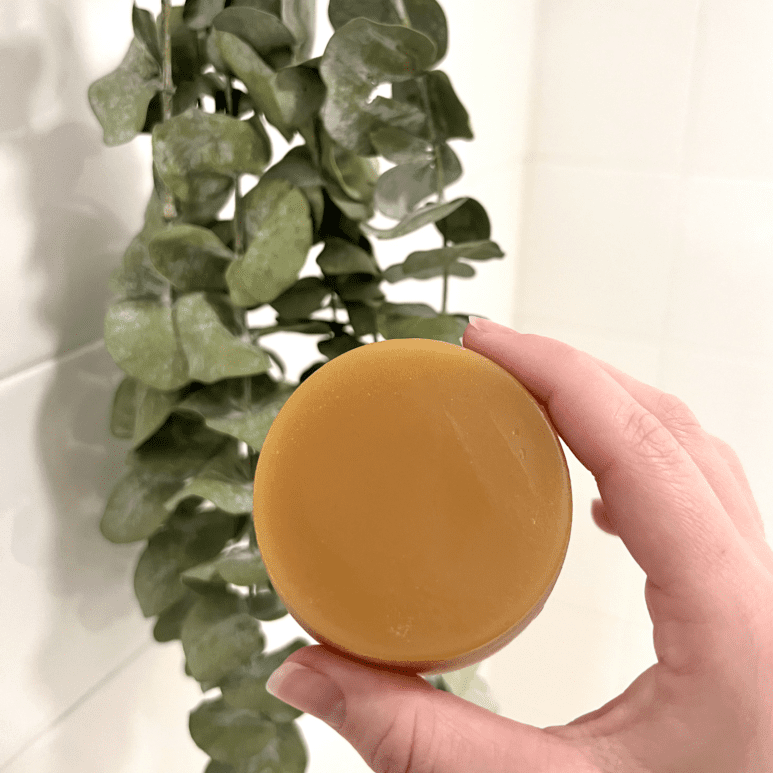 A hand holding a full-sized conditioner bar. In the faded background is a white tile shower with eucalyptus.